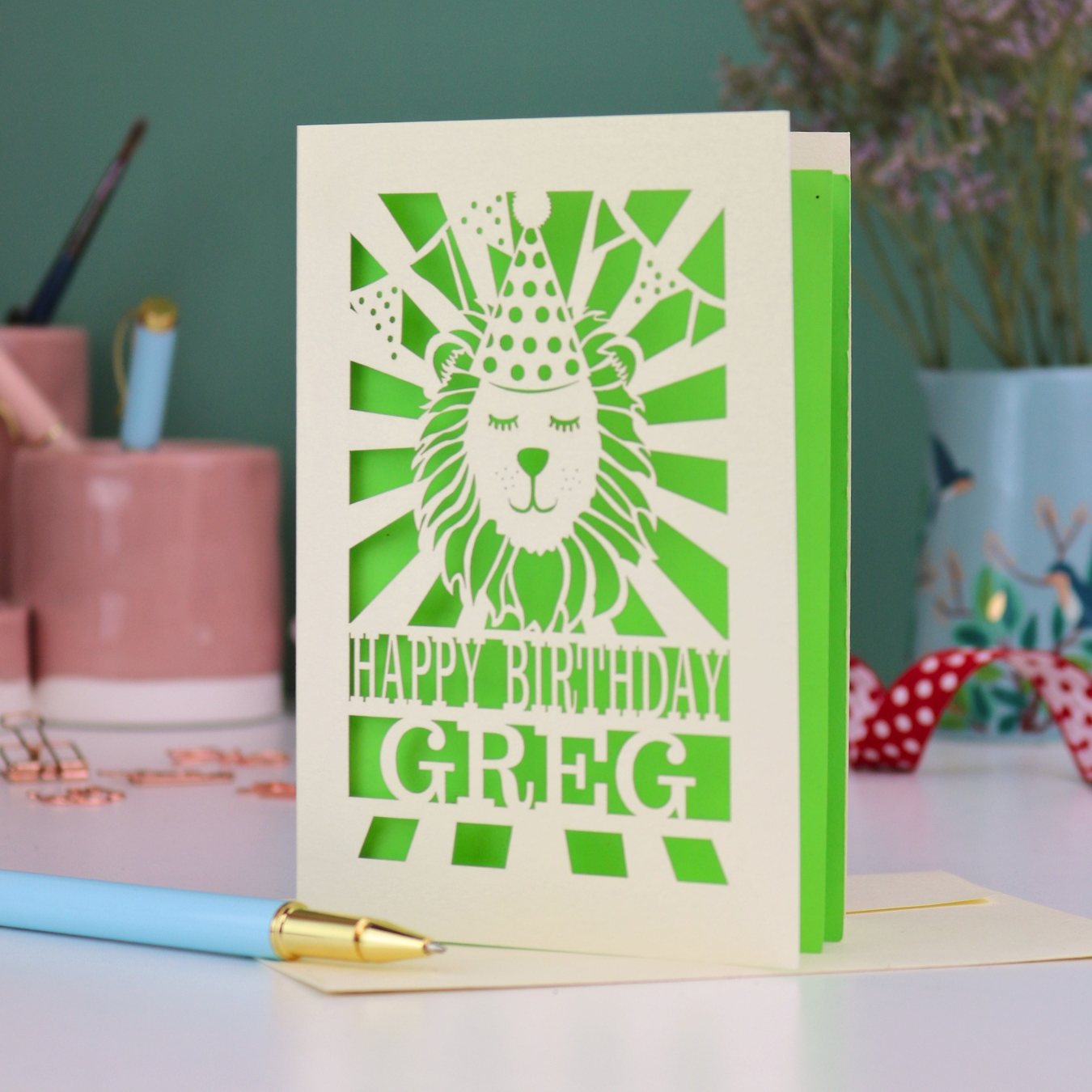 A personalised birthday card with laser cut lion in a party hat design and personalised text "Happy Birthday Greg", on a white desk with pen and stationery items
