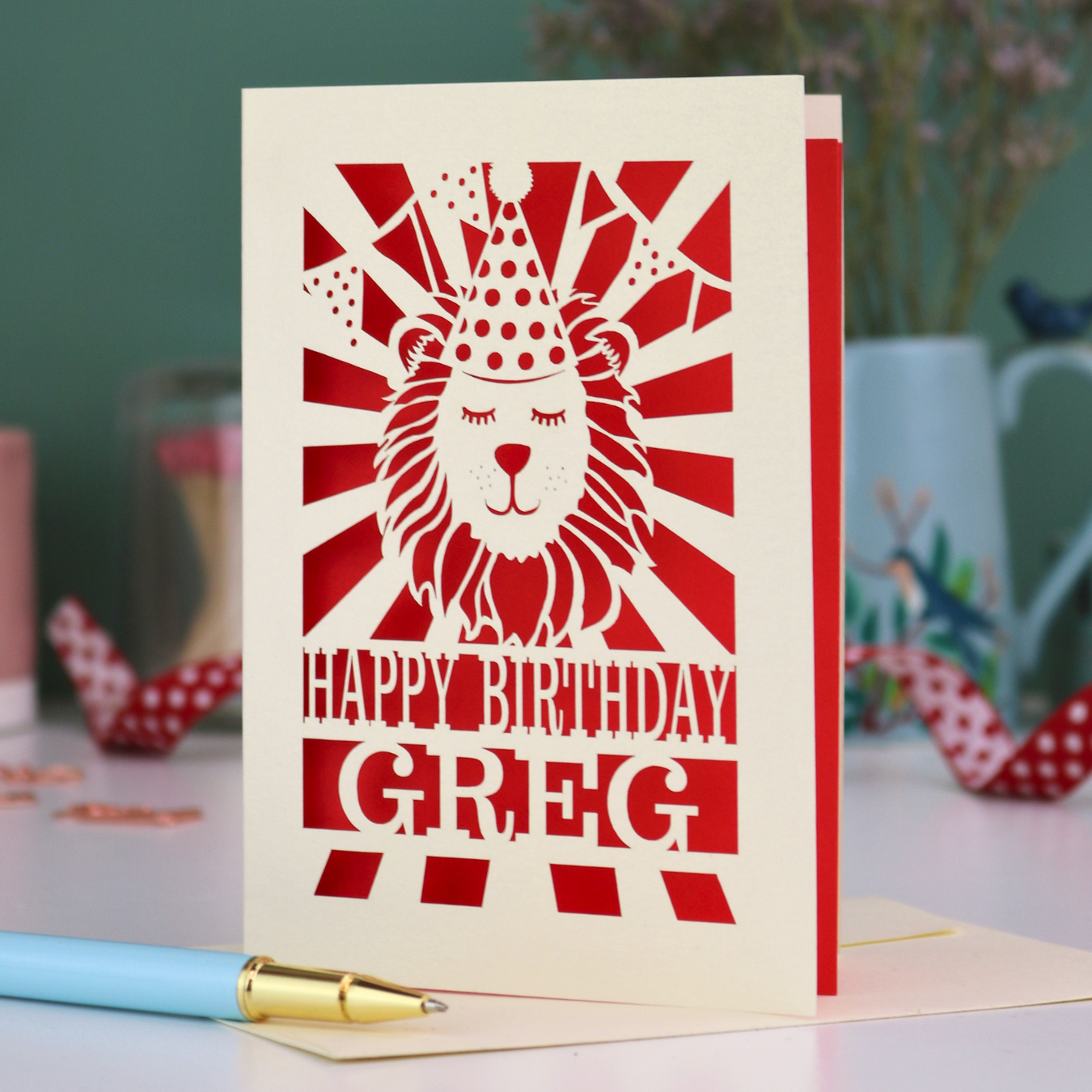 A personalised birthday card with laser cut lion in a party hat design and personalised text "Happy Birthday Greg", on a white desk with pen and stationery items