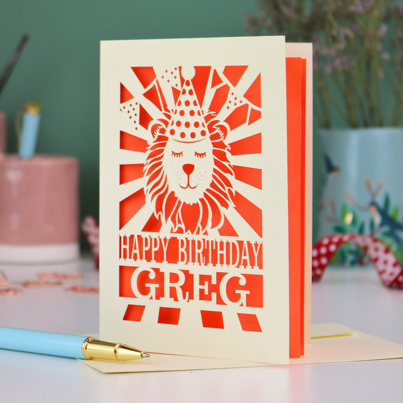 A personalised birthday card with laser cut lion in a party hat design and personalised text "Happy Birthday Greg", on a white desk with pen and stationery items