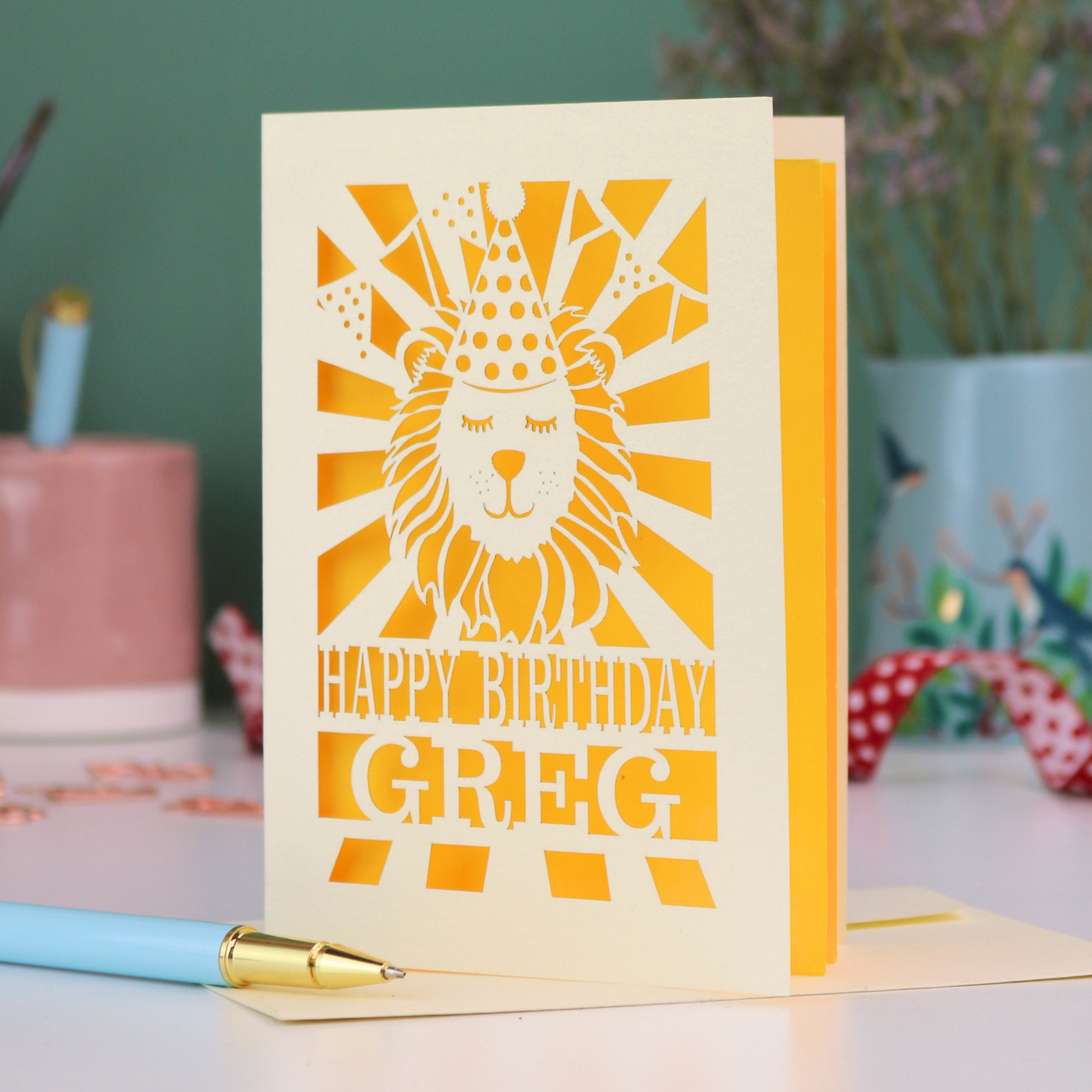 A personalised birthday card with laser cut lion in a party hat design and personalised text "Happy Birthday Greg", on a white desk with pen and stationery items