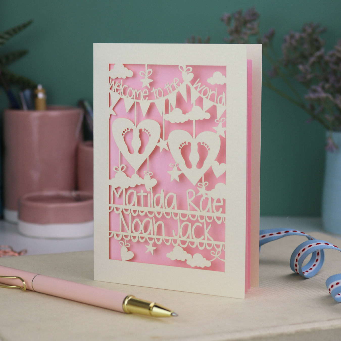 A laser cut twins card with 'Welcome to the World', personalised with the twins' names on two lines of text and with two pairs of baby feet cut out from two heart shapes. 
