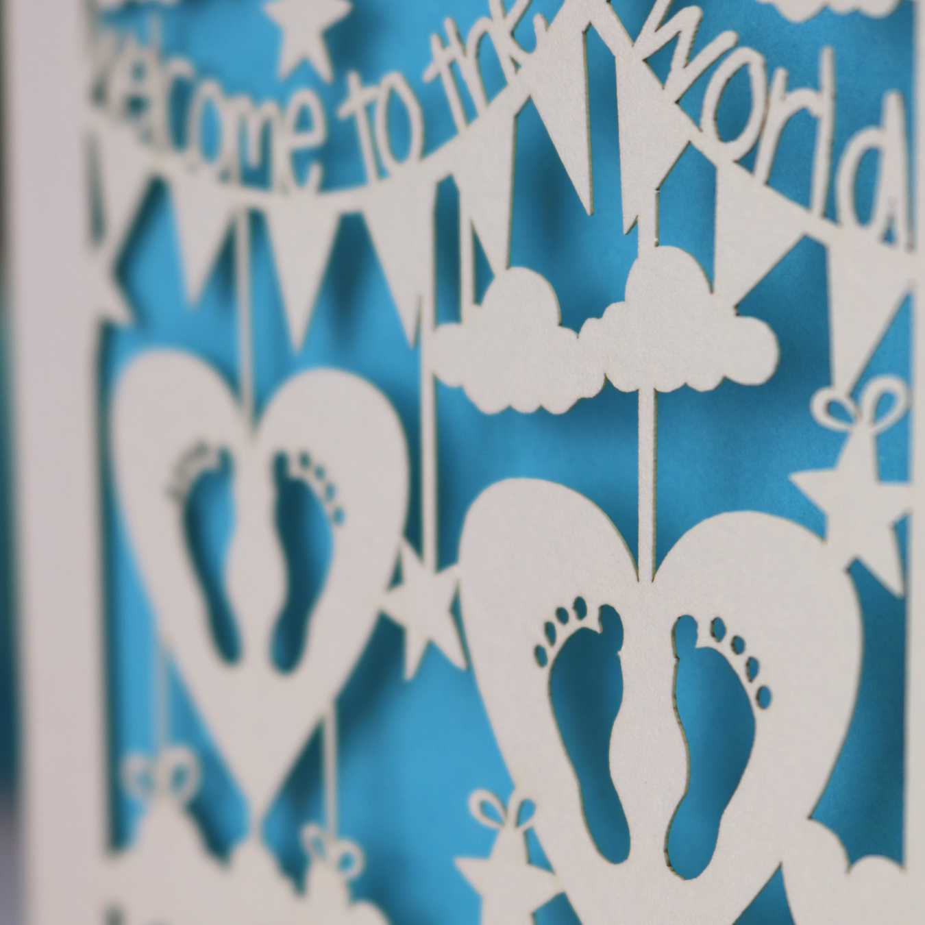 A laser cut twins card with 'Welcome to the World', personalised with the twins' names on two lines of text and with two pairs of baby feet cut out from two heart shapes. 