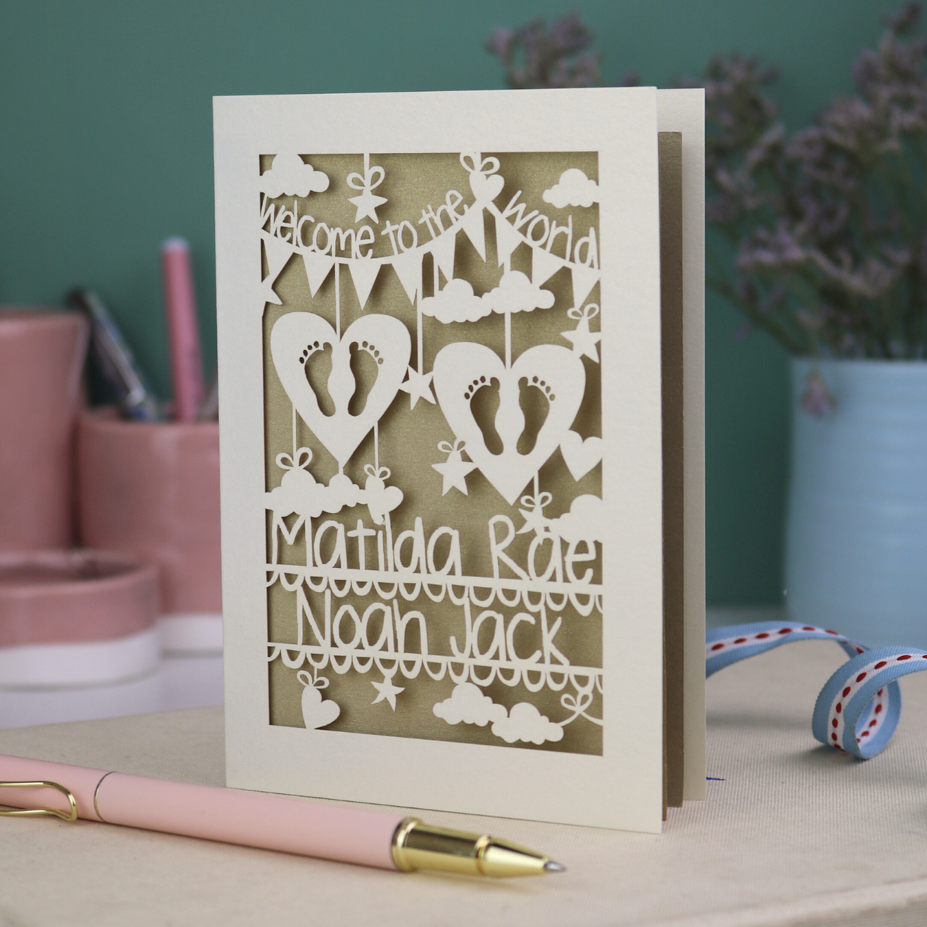 A laser cut twins card with 'Welcome to the World', personalised with the twins' names on two lines of text and with two pairs of baby feet cut out from two heart shapes. 