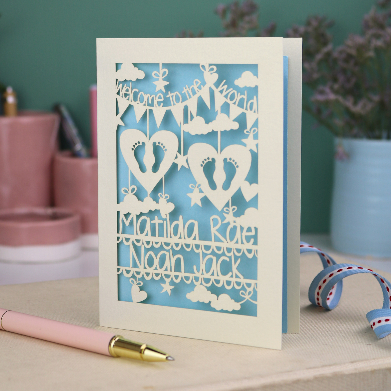 A laser cut twins card with 'Welcome to the World', personalised with the twins' names on two lines of text and with two pairs of baby feet cut out from two heart shapes. 