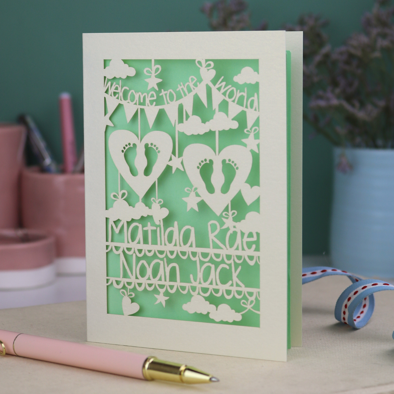A laser cut twins card with 'Welcome to the World', personalised with the twins' names on two lines of text and with two pairs of baby feet cut out from two heart shapes. 