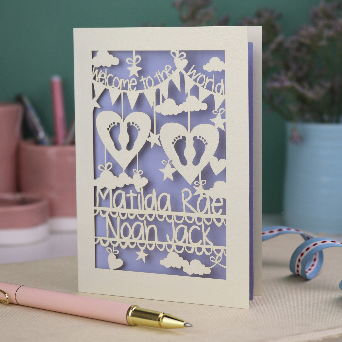 A laser cut twins card with 'Welcome to the World', personalised with the twins' names on two lines of text and with two pairs of baby feet cut out from two heart shapes. 