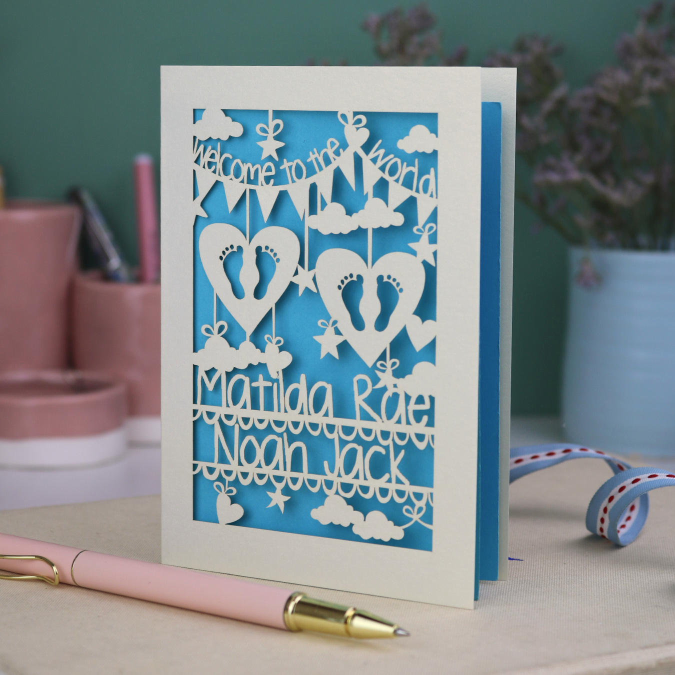 A laser cut twins card with 'Welcome to the World', personalised with the twins' names on two lines of text and with two pairs of baby feet cut out from two heart shapes. 