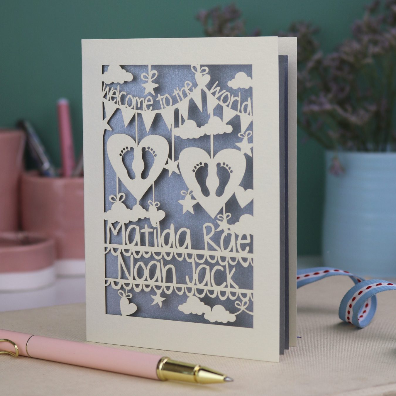 A laser cut twins card with 'Welcome to the World', personalised with the twins' names on two lines of text and with two pairs of baby feet cut out from two heart shapes. 