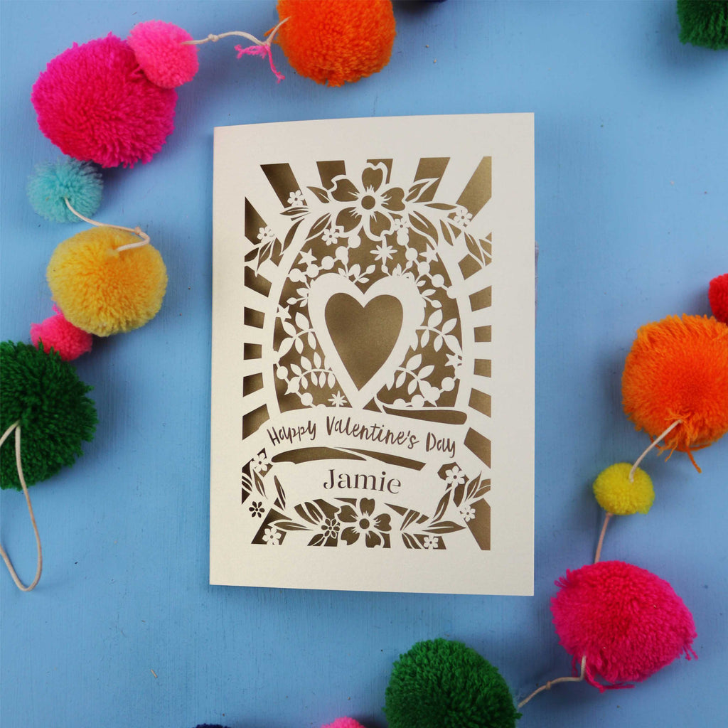 Decorative cut out Valentine's card with floral heart design on a blue background with colourful pom-poms.