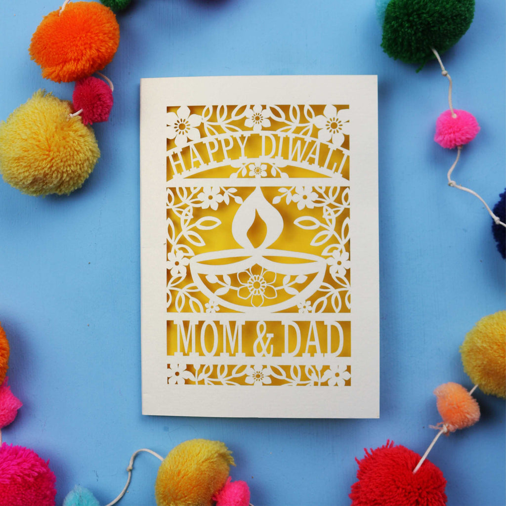 Decorative Diwali card with 'Happy Diwali' and 'Mom & Dad' text on a blue background with colourful pom-poms.