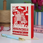 A laser cut 1st birthday card personalised with a name and age, and featuring a cut out pirate ship design in cream on a red background