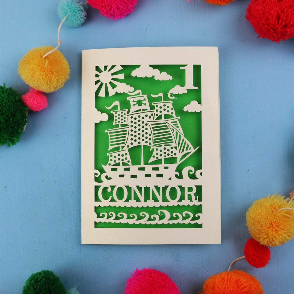 Decorative 1st Birthday card with ship design and 'CONNOR' text on a blue background with colourful pom-poms.