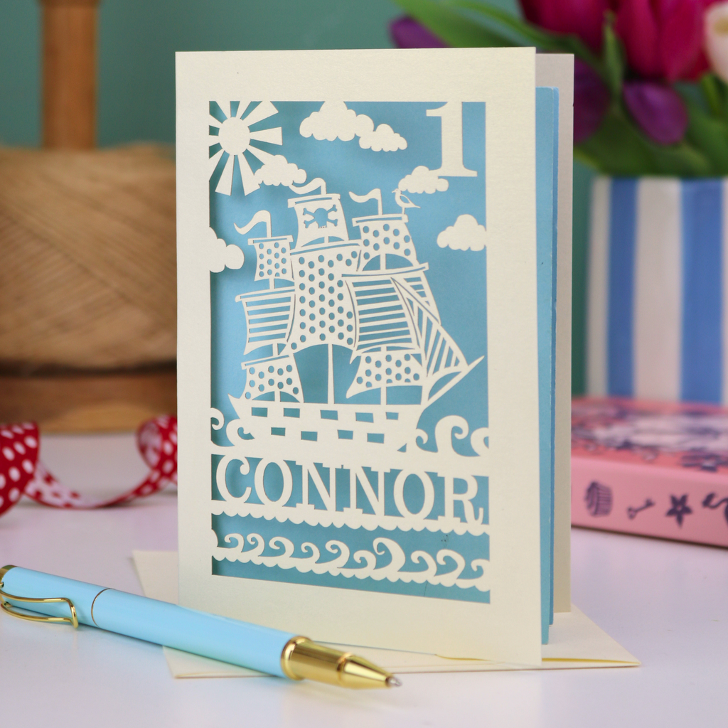 A laser cut 1st birthday card personalised with a name and age, and featuring a cut out pirate ship design in cream on a light blue background