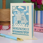 A laser cut 1st birthday card personalised with a name and age, and featuring a cut out pirate ship design in cream on a light blue background