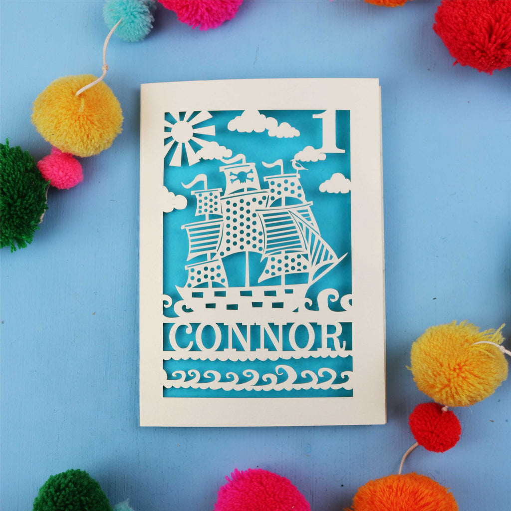 Decorative 1st Birthday card with ship design and 'CONNOR' text on a blue background with colourful pom-poms.