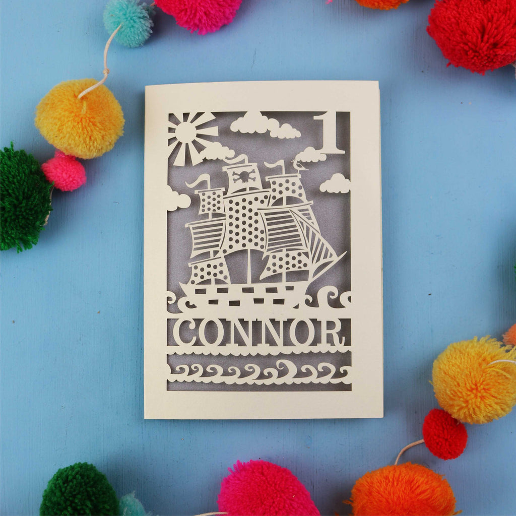 Decorative 1st Birthday card with ship design and 'CONNOR' text on a blue background with colourful pom-poms.