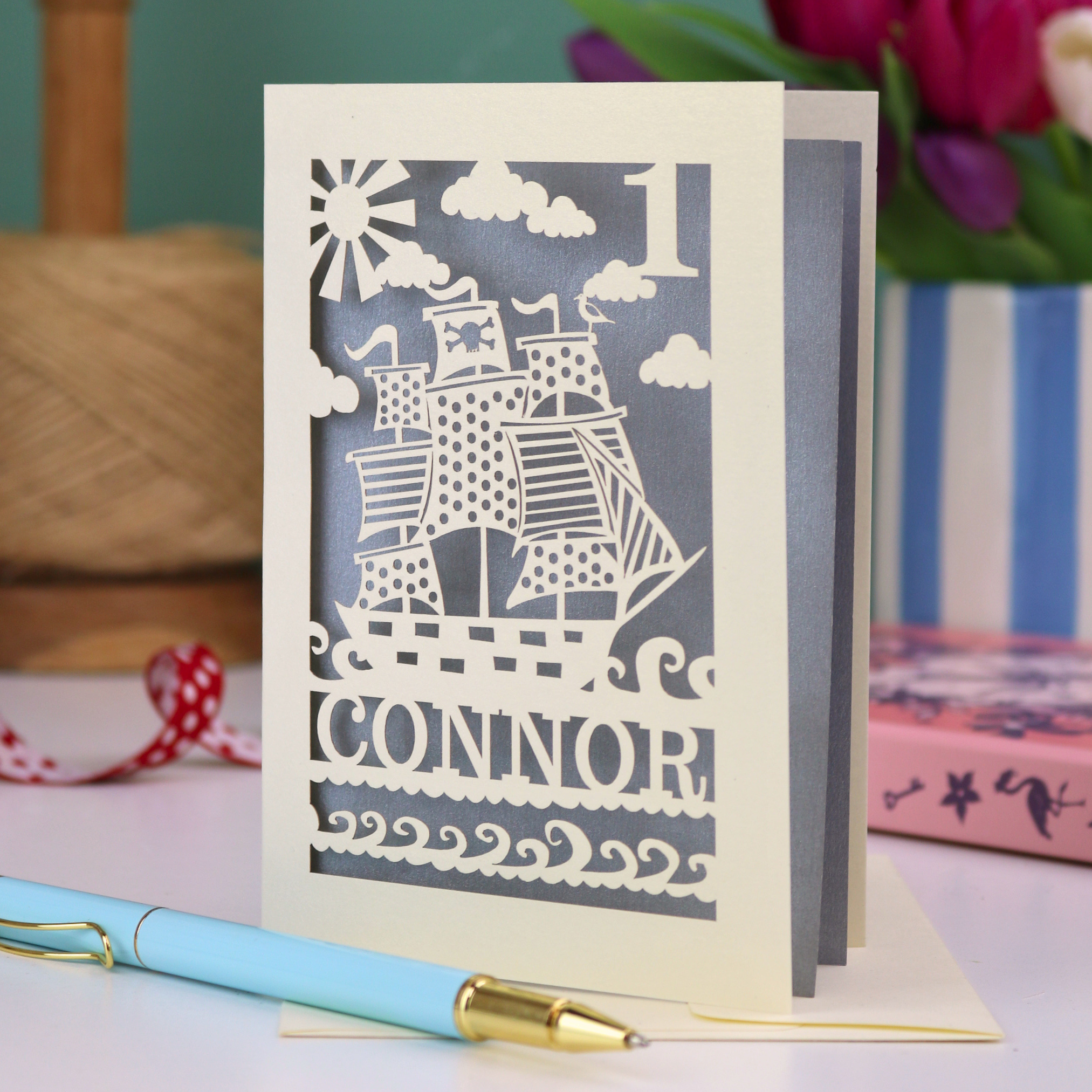 A laser cut 1st birthday card personalised with a name and age, and featuring a cut out pirate ship design in cream on a silver background