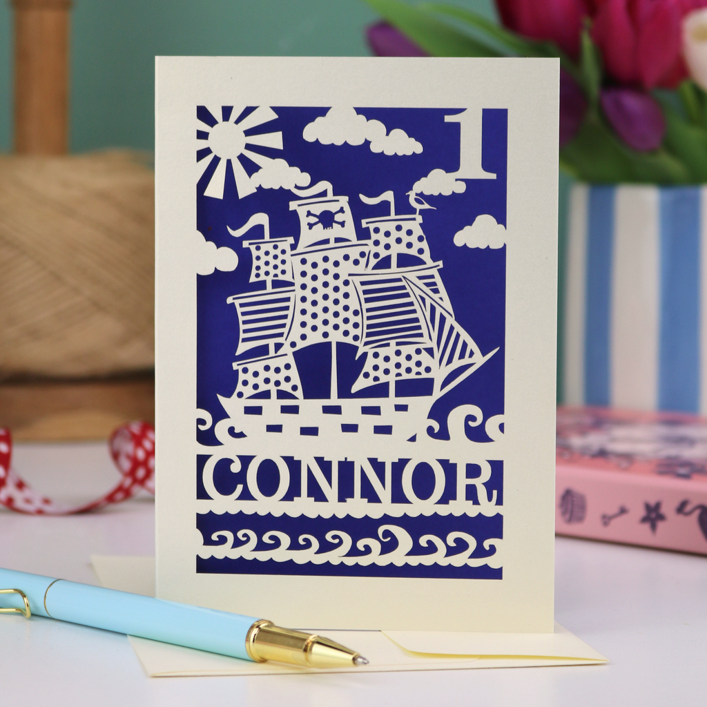 A laser cut 1st birthday card personalised with a name and age, and featuring a cut out pirate ship design in cream on a violet background