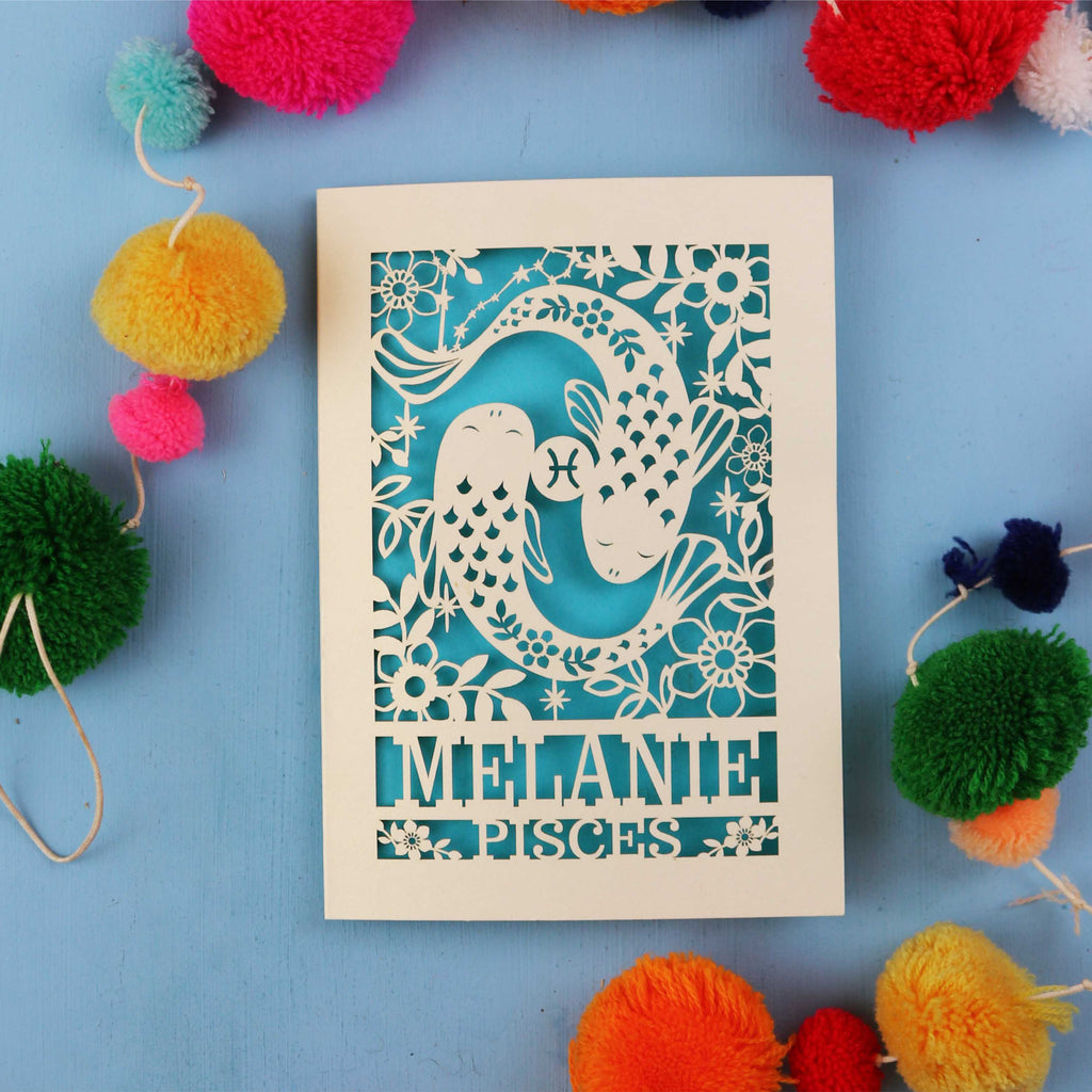 Decorative birthday card with cut-out design and 'Melanie Pisces' text on a blue background with colourful pom-poms.