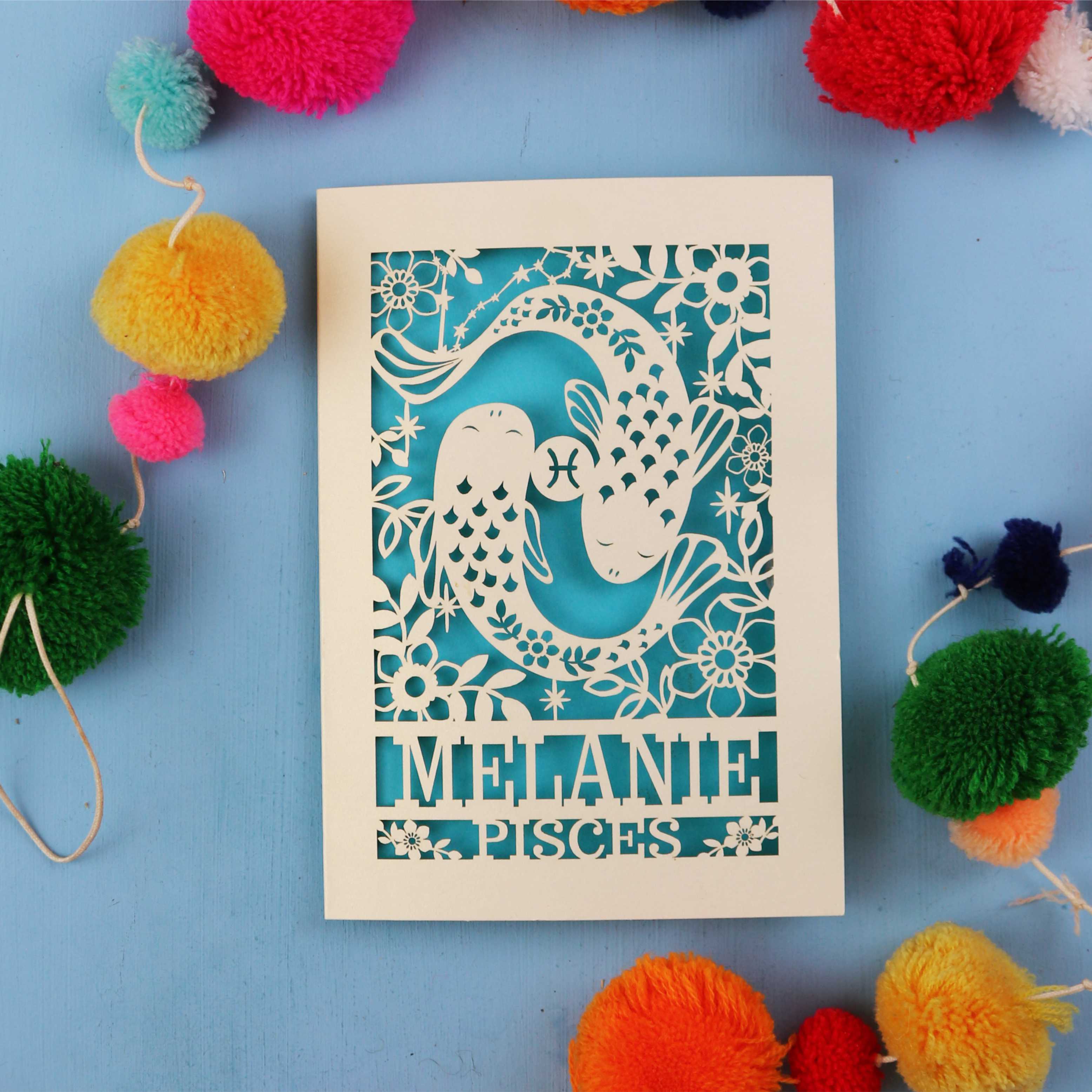 Decorative birthday card with cut-out design and 'Melanie Pisces' text on a blue background with colourful pom-poms.