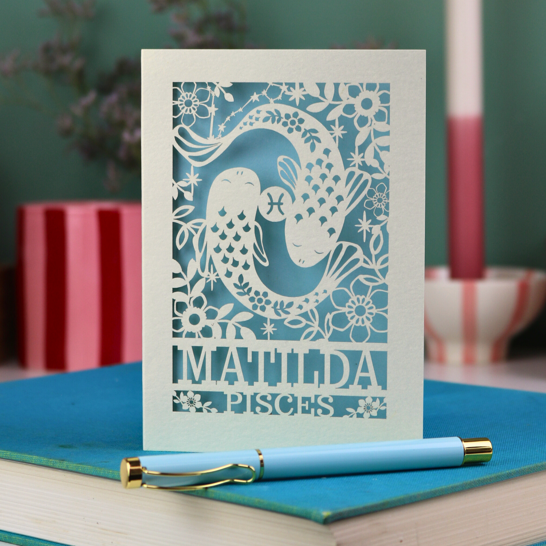 Decorative Pisces birthday card with 'Matilda' and a laser cut Pisces design on a blue surface with a pen.