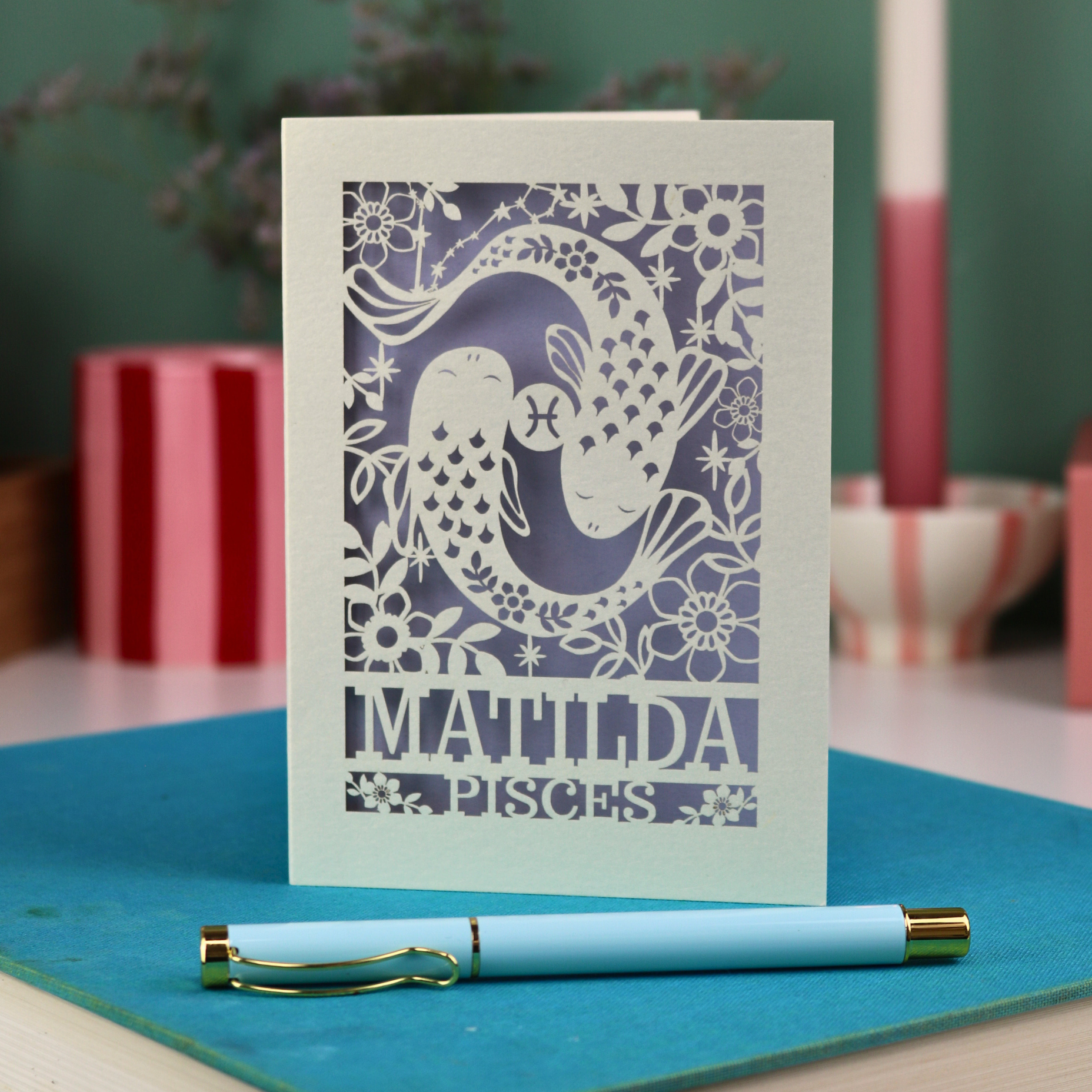 Decorative Pisces birthday card with 'Matilda' and a laser cut Pisces design on a blue surface with a pen.