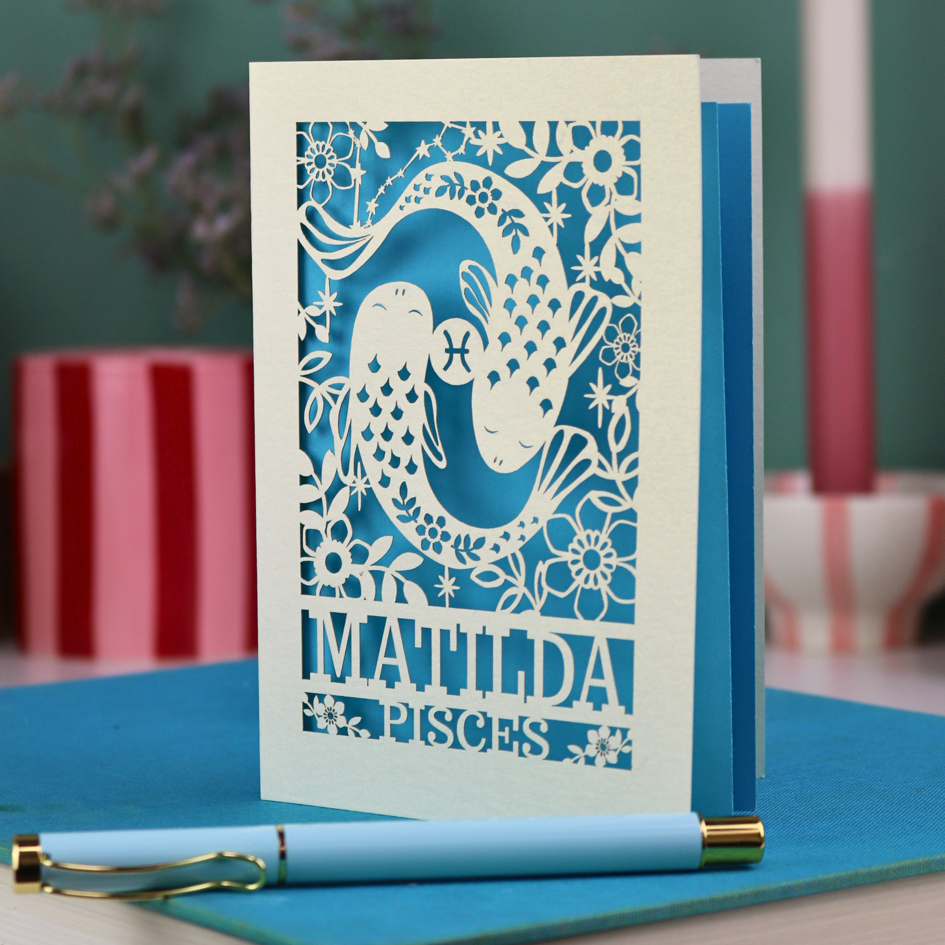 Decorative Pisces birthday card with 'Matilda' and a laser cut Pisces design on a blue surface with a pen.