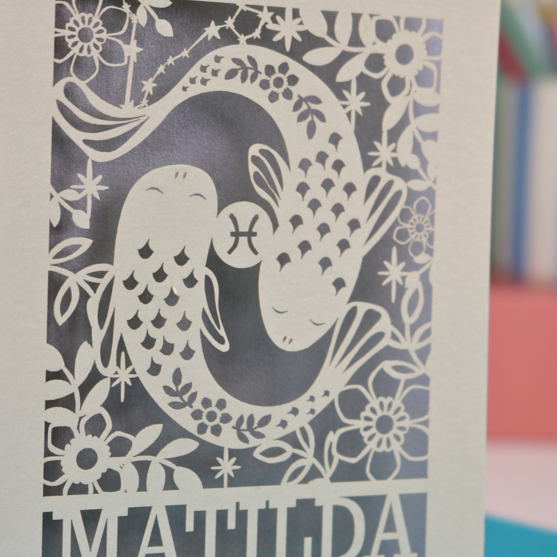 Decorative paper cut out of two fish with floral elements on a blue background, featuring the name 'MATILDA'.