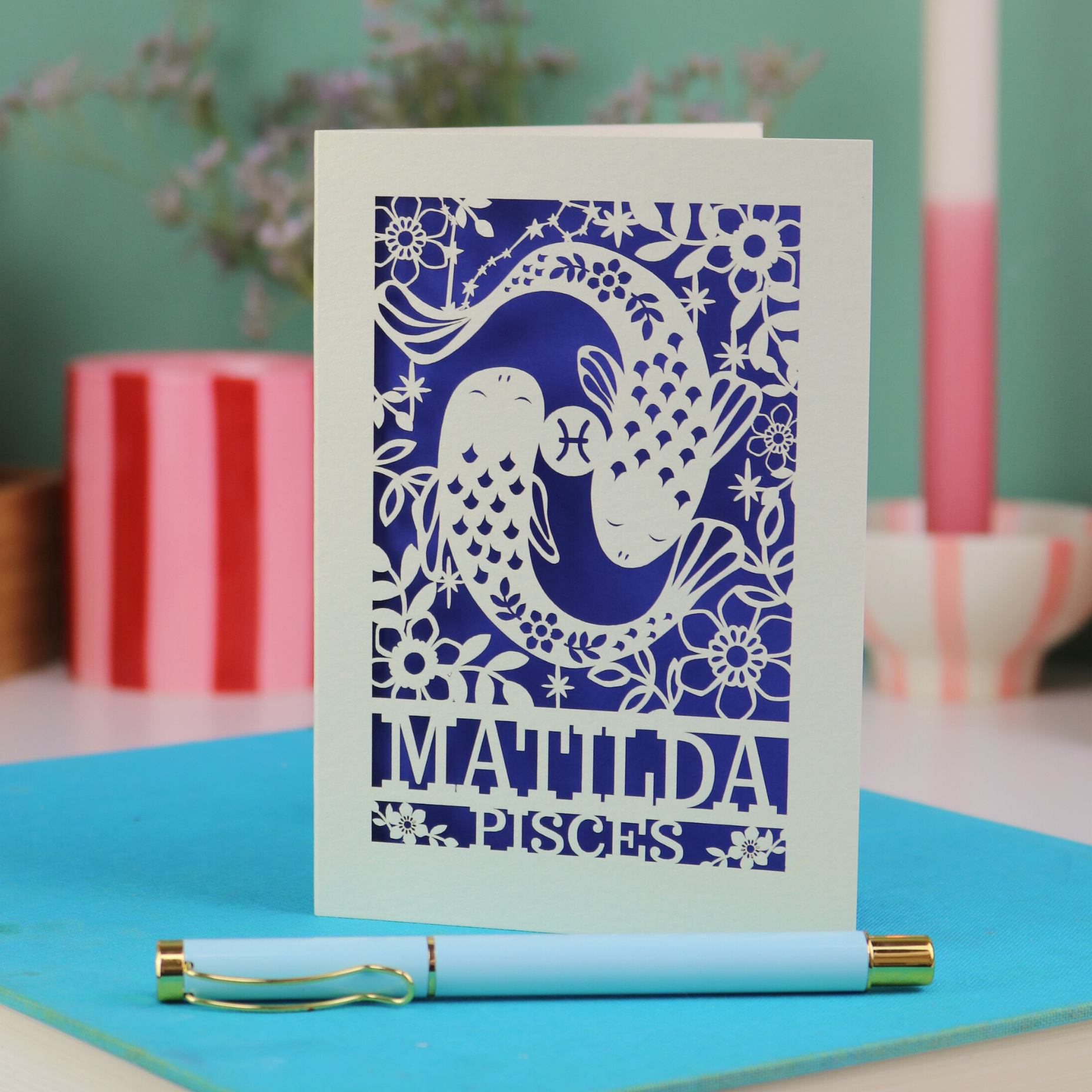 Decorative Pisces birthday card with 'Matilda' and a laser cut Pisces design on a blue surface with a pen.