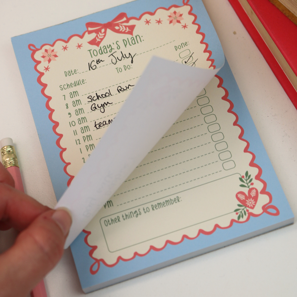 A close-up of a light blue notepad with a red scalloped border and floral corner details. A hand is peeling back a page to show the writing surface underneath.
