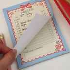 A close-up of a light blue notepad with a red scalloped border and floral corner details. A hand is peeling back a page to show the writing surface underneath.
