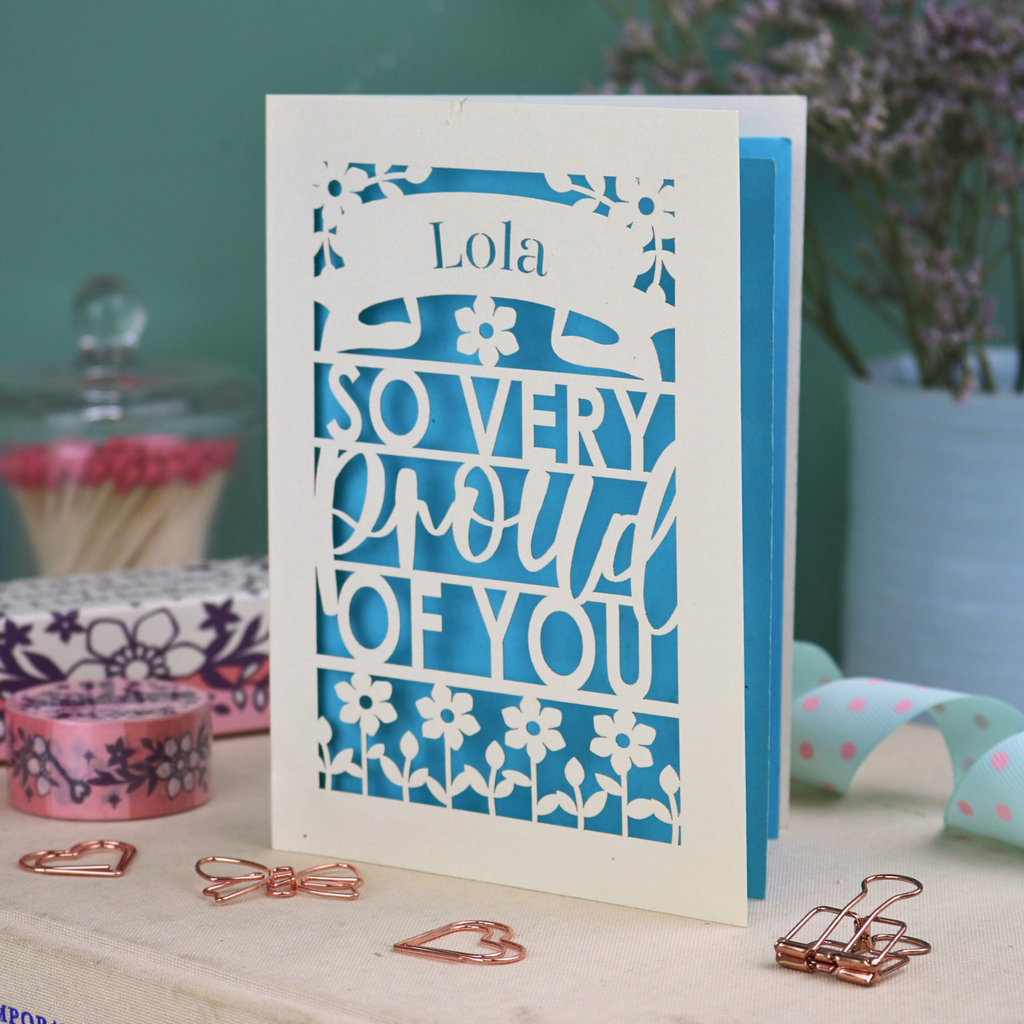 A personalised cut out congratulations card with 'Lola, so very proud of you' text on a decorative surface with flowers and stationery items.
