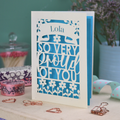 A personalised cut out congratulations card with 'Lola, so very proud of you' text on a decorative surface with flowers and stationery items.