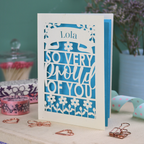 A personalised cut out congratulations card with 'Lola, so very proud of you' text on a decorative surface with flowers and stationery items.
