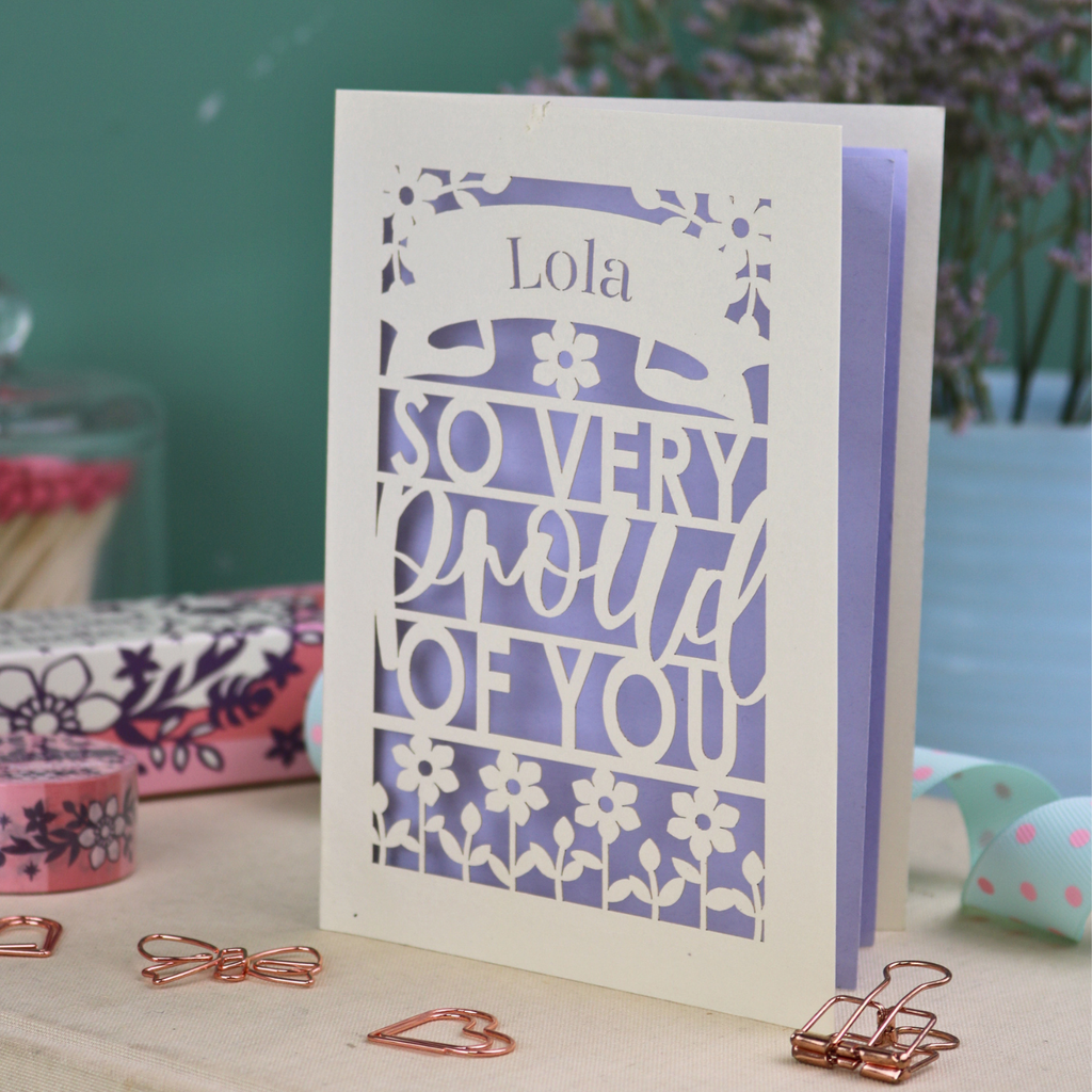 A personalised laser cut pride card with 'Lola, so very proud of you' text on a decorative surface with flowers and stationery items.