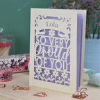 A personalised laser cut pride card with 'Lola, so very proud of you' text on a decorative surface with flowers and stationery items.
