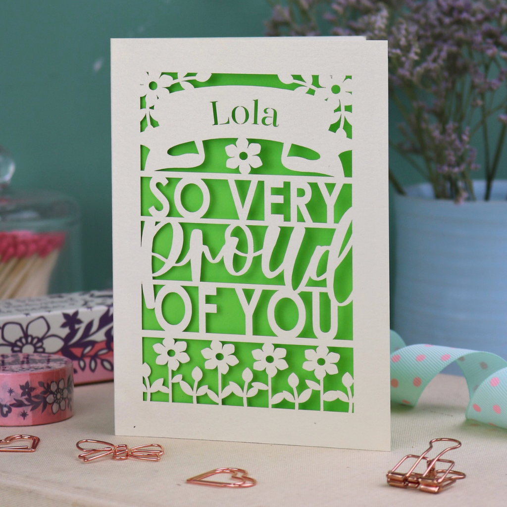 A personalised congratulations card with 'Lola, so very proud of you' text on a decorative surface with flowers and stationery items.