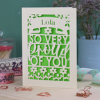 A personalised congratulations card with 'Lola, so very proud of you' text on a decorative surface with flowers and stationery items.