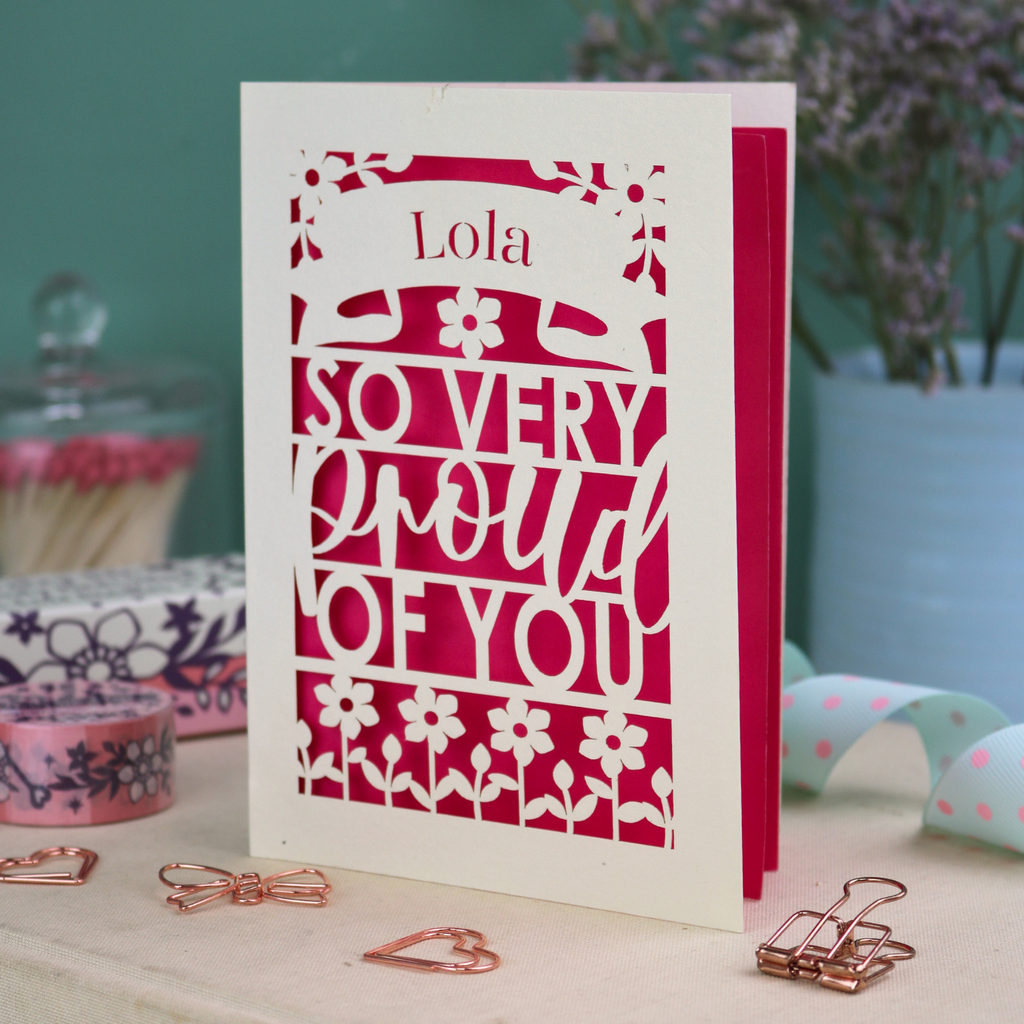 A personalised congratulations on your success card with 'Lola, so very proud of you' text on a decorative surface with flowers and stationery items.