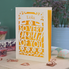 A personalised laser cut exam pass card with 'Lola, so very proud of you' text on a decorative surface with flowers and stationery items.