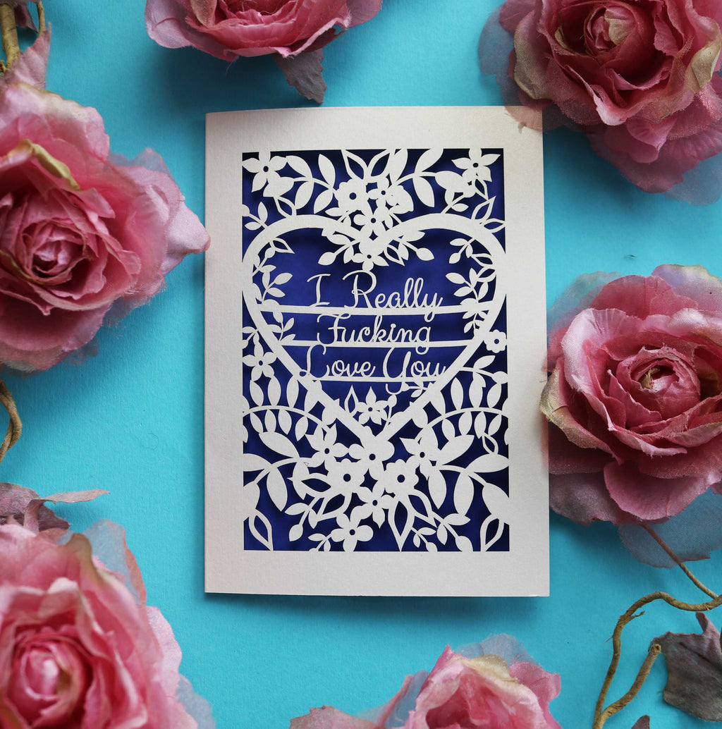 A unusual and unique rude valentines card that is laser cut. Design is cut out from cream card to show purple paper behind. Cut out design reads "I really fucking love you" inside a heart shape, surrounded by floral designs.