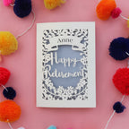 A personalised laser cut retirement card. Card has a banner with a name and the words "Happy Retirement" in a script font, surrounded but flowers and leaves.