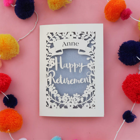 A personalised laser cut retirement card. Card has a banner with a name and the words "Happy Retirement" in a script font, surrounded but flowers and leaves.