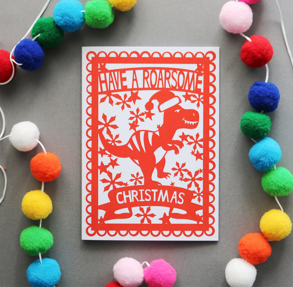 A Christmas card with a red dinosaur illustration surrounded by festive snowflakes, with the text 'HAVE A ROARSOME CHRISTMAS' across the top. The card is displayed on a grey background with a colourful pom-pom garland around it.