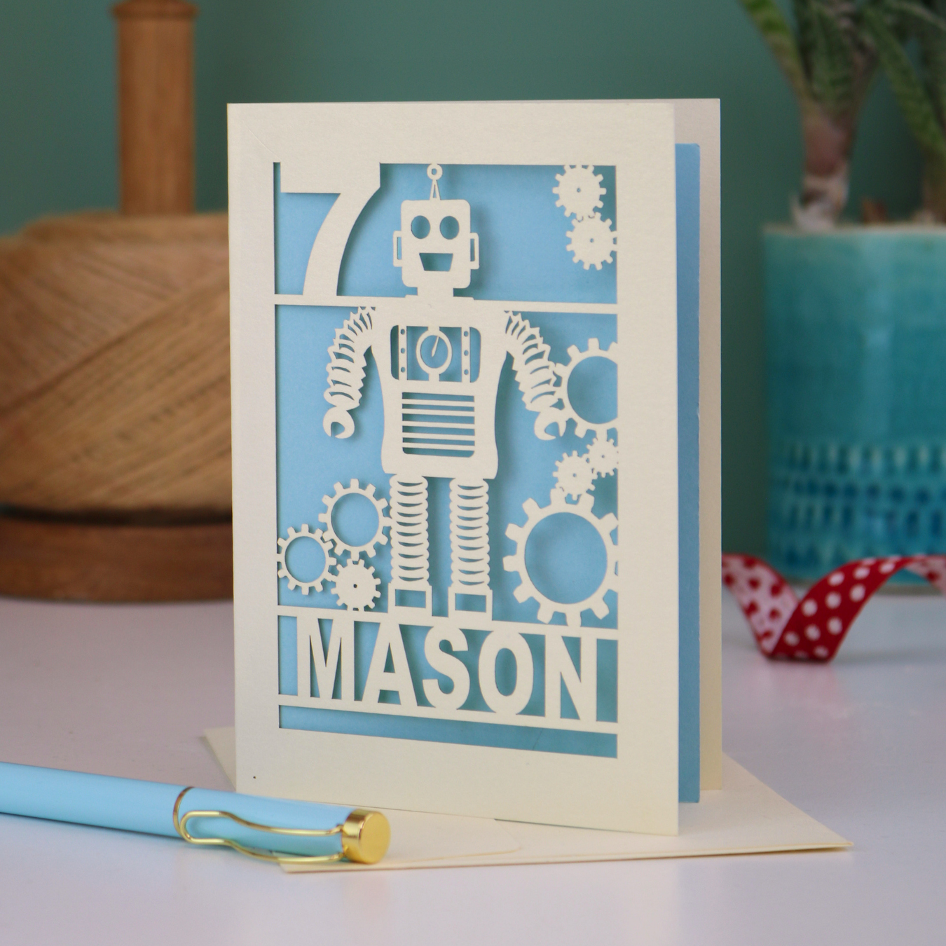 A cream and light blue laser cut birthday card with robot design and personalised with the age and name of the recipient