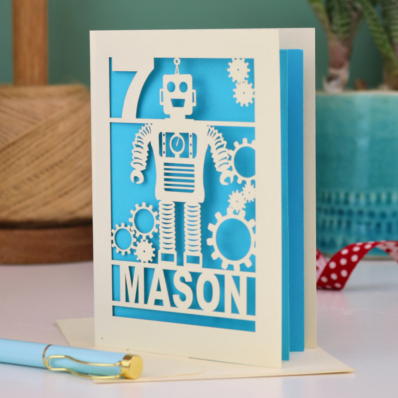 A cream and peacock blue laser cut birthday card with robot design and personalised with the age and name of the recipient