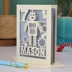 A cream and silver laser cut birthday card with robot design and personalised with the age and name of the recipient