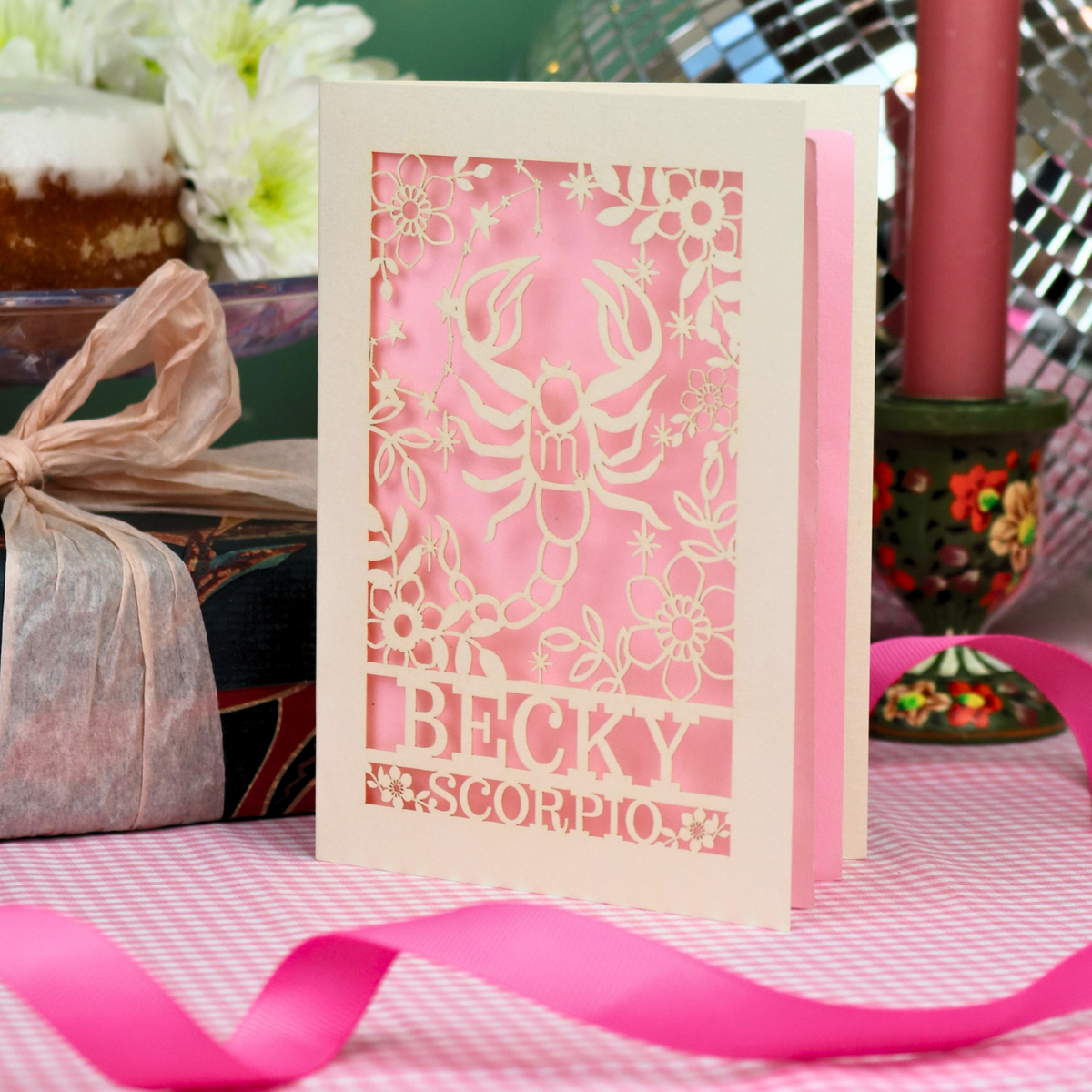 A handmade laser-cut greeting card featuring a cream Scorpio zodiac design over a pastel pink background, with the name 'BECKY' and 'SCORPIO'. The card is styled on a pink-and-white checked tablecloth with a pink ribbon, surrounded by party decorations.