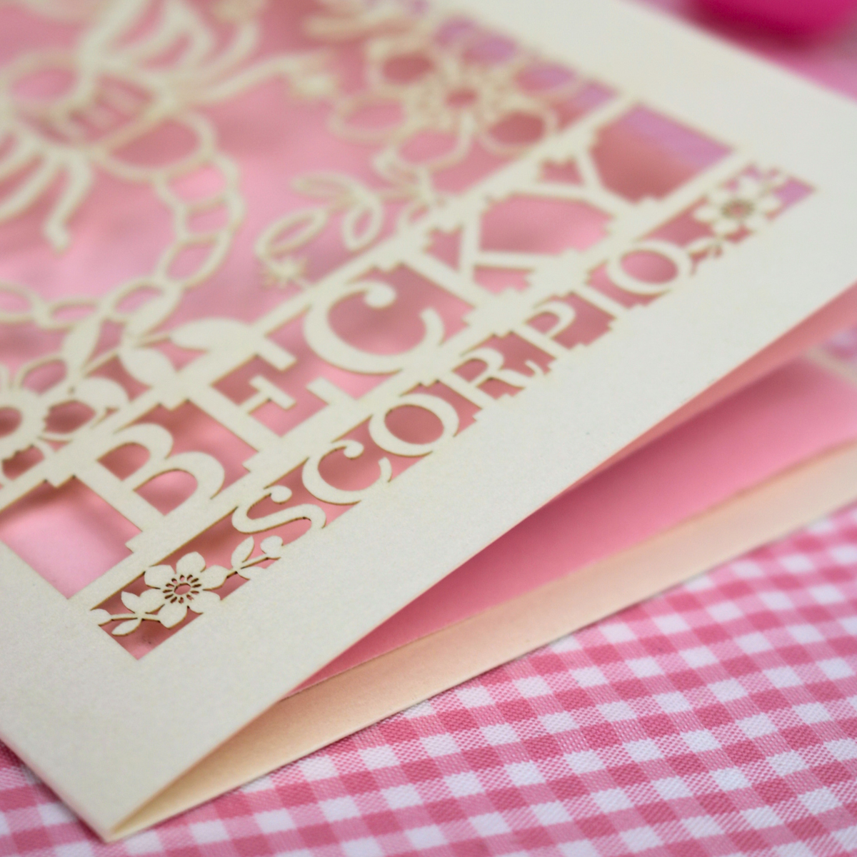 A close-up of a laser-cut cream card with floral and script details, including the words 'BECKY' and 'SCORPIO', layered over pink cardstock on a pink-and-white gingham surface.