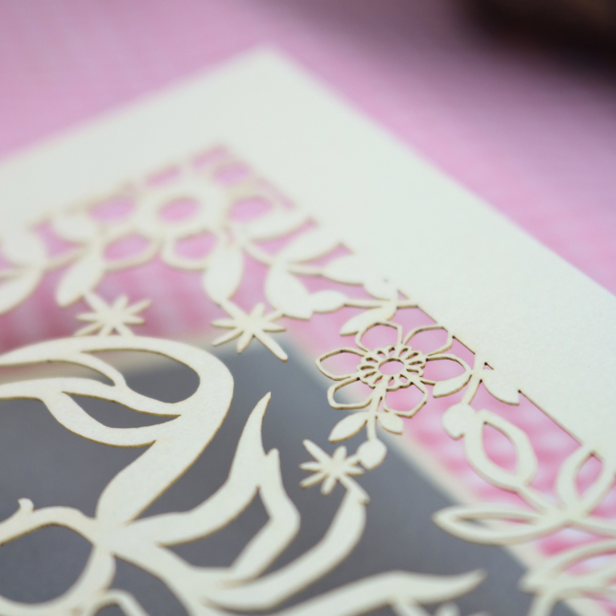 A close-up of a laser-cut decorative card with floral and abstract cut out patterns, showing a white card over a pink and silver background.